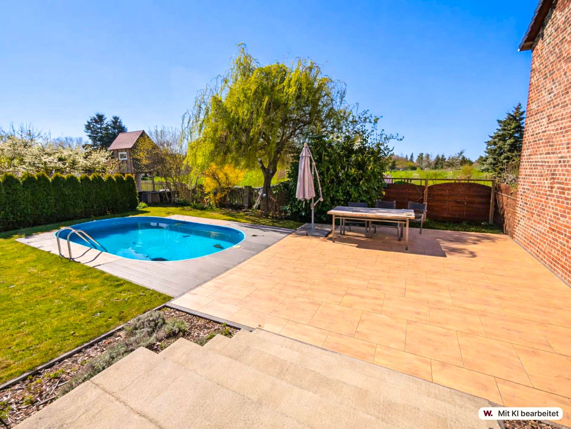 Backyard patio with blue pool, outdoor table and umbrella, and brick house wall on the right under a clear blue sky.