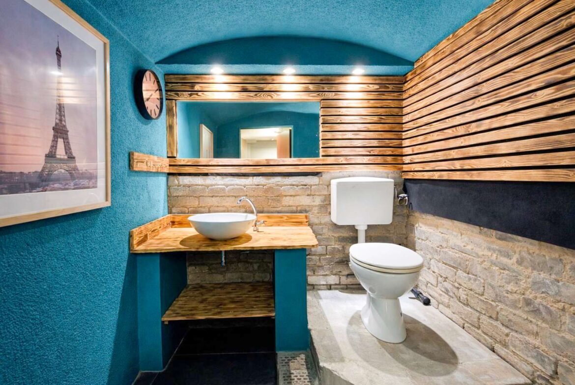 Teal-walled bathroom with a wooden sink counter, vessel basin, large mirror, brick lower wall, and a white toilet.