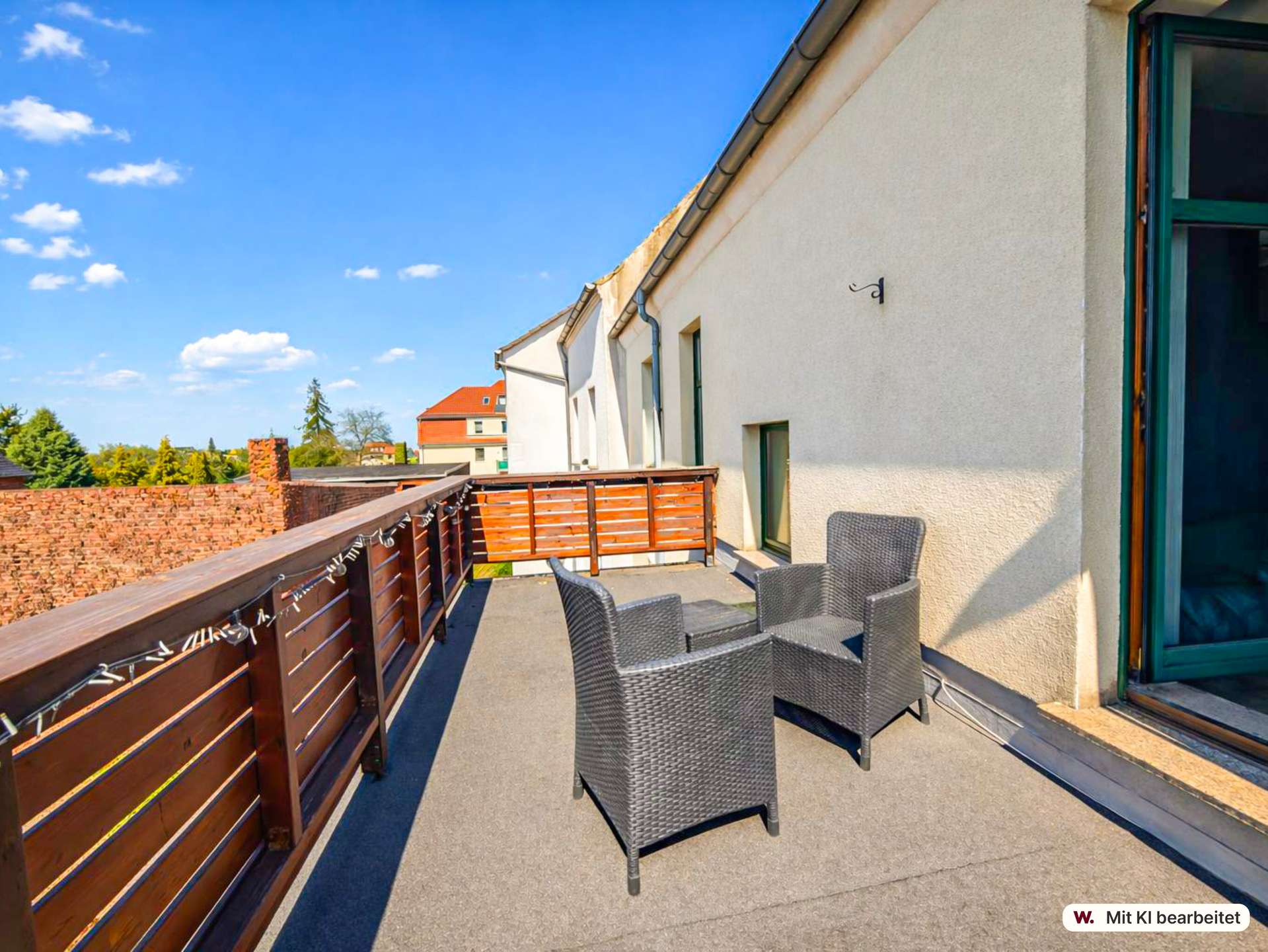 Sunny rooftop balcony with brown wooden railing and gray wicker chairs facing outward, beige building wall on the right and clear blue sky above.