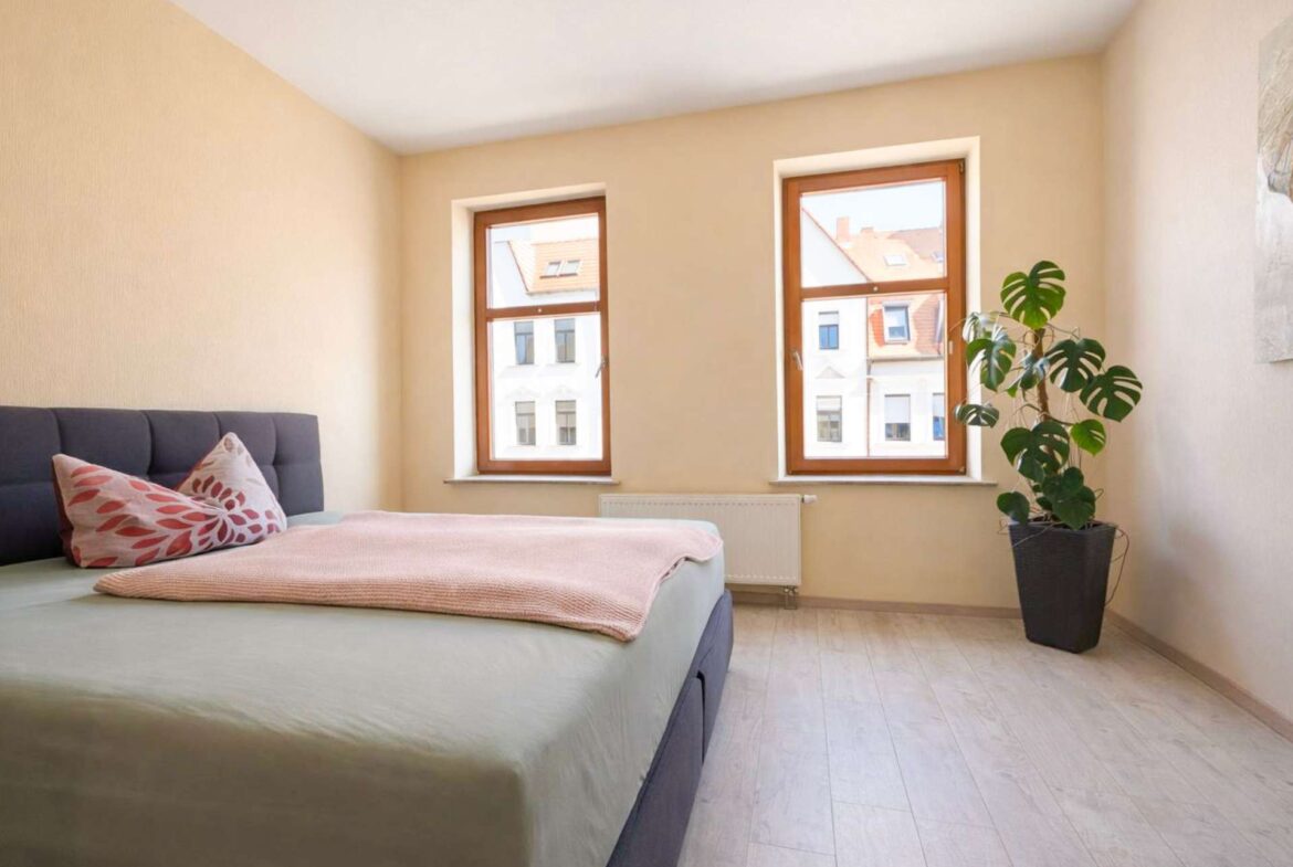 Bedroom with two wooden-framed windows, a potted Monstera plant, and a neatly made bed with pink cushions and soft pink throw on light wood floors.