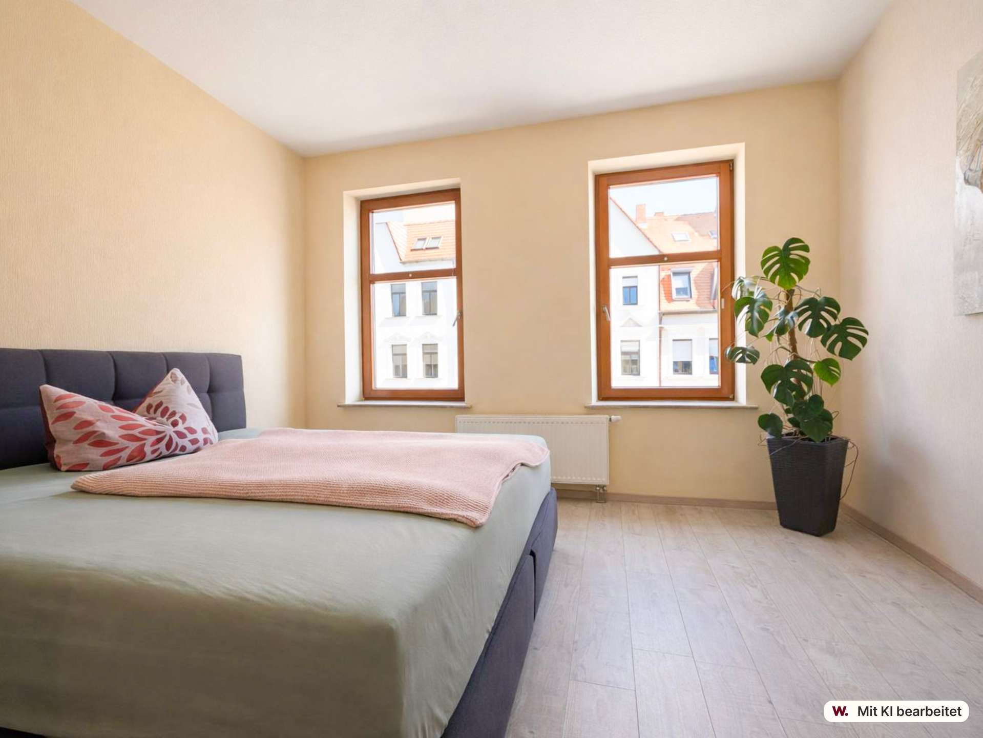 Bedroom with two wooden-framed windows, a potted Monstera plant, and a neatly made bed with pink cushions and soft pink throw on light wood floors.
