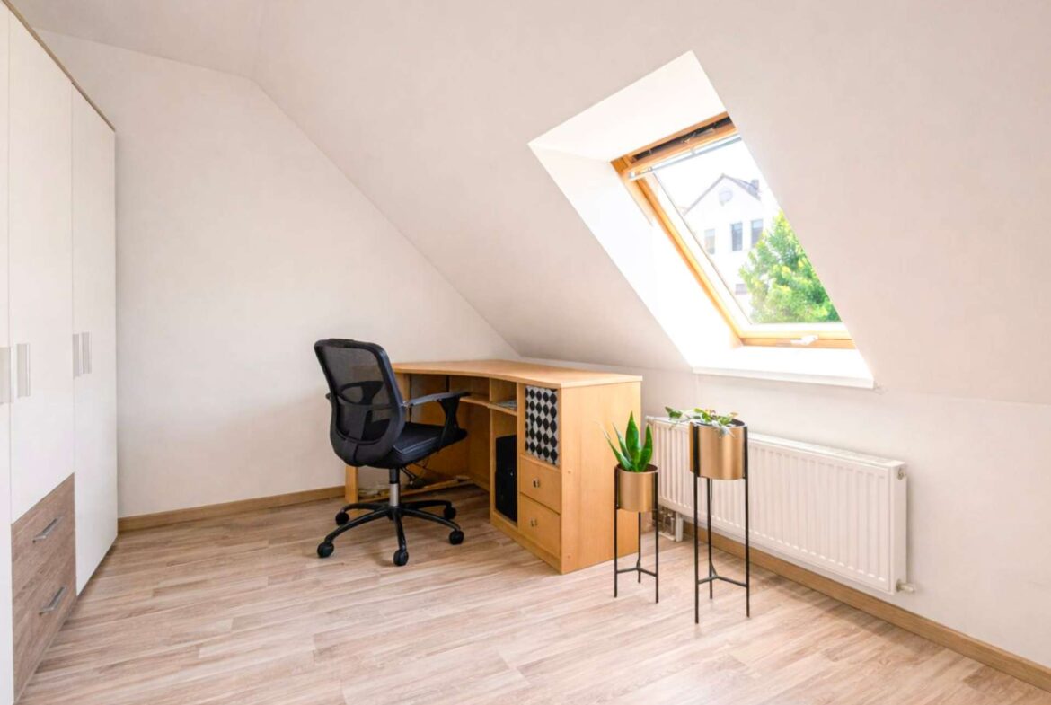 Home office under a sloped ceiling with a wooden desk, black ergonomic chair, and a skylight window.
