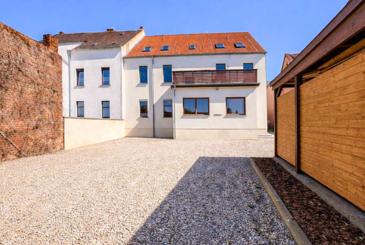 Courtyard with pebble ground, white multi-story building, red-tiled roof, and a wooden fence on the right under a blue sky.