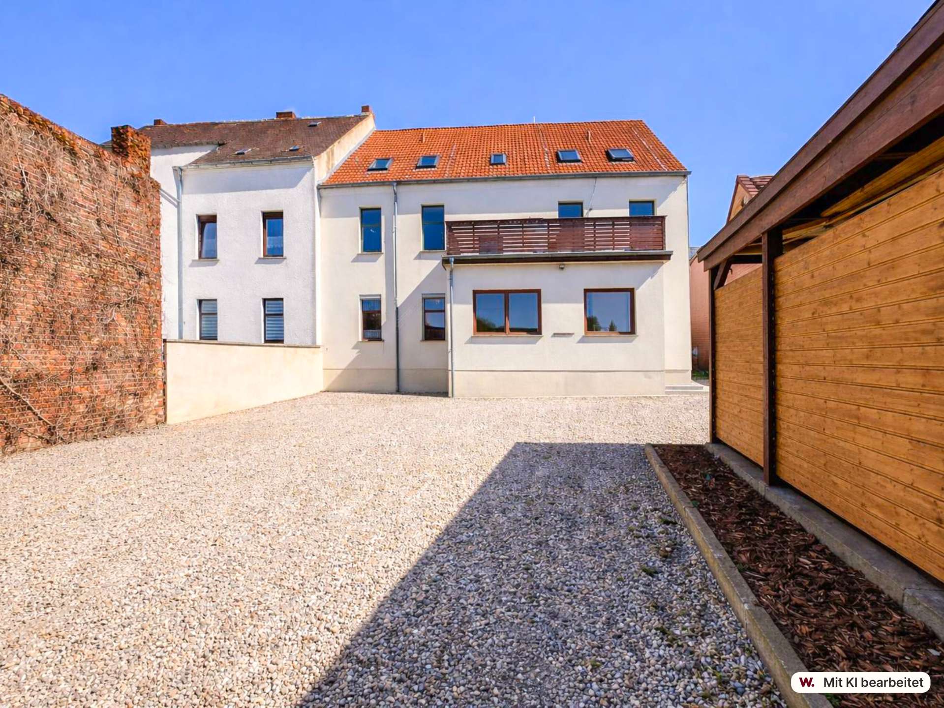 Courtyard with pebble ground, white multi-story building, red-tiled roof, and a wooden fence on the right under a blue sky.
