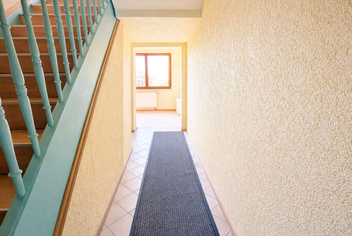 Indoor hallway and staircase with a turquoise railing on the left, beige textured walls, and a dark gray rug along tiled floor toward a sunlit doorway at the end.