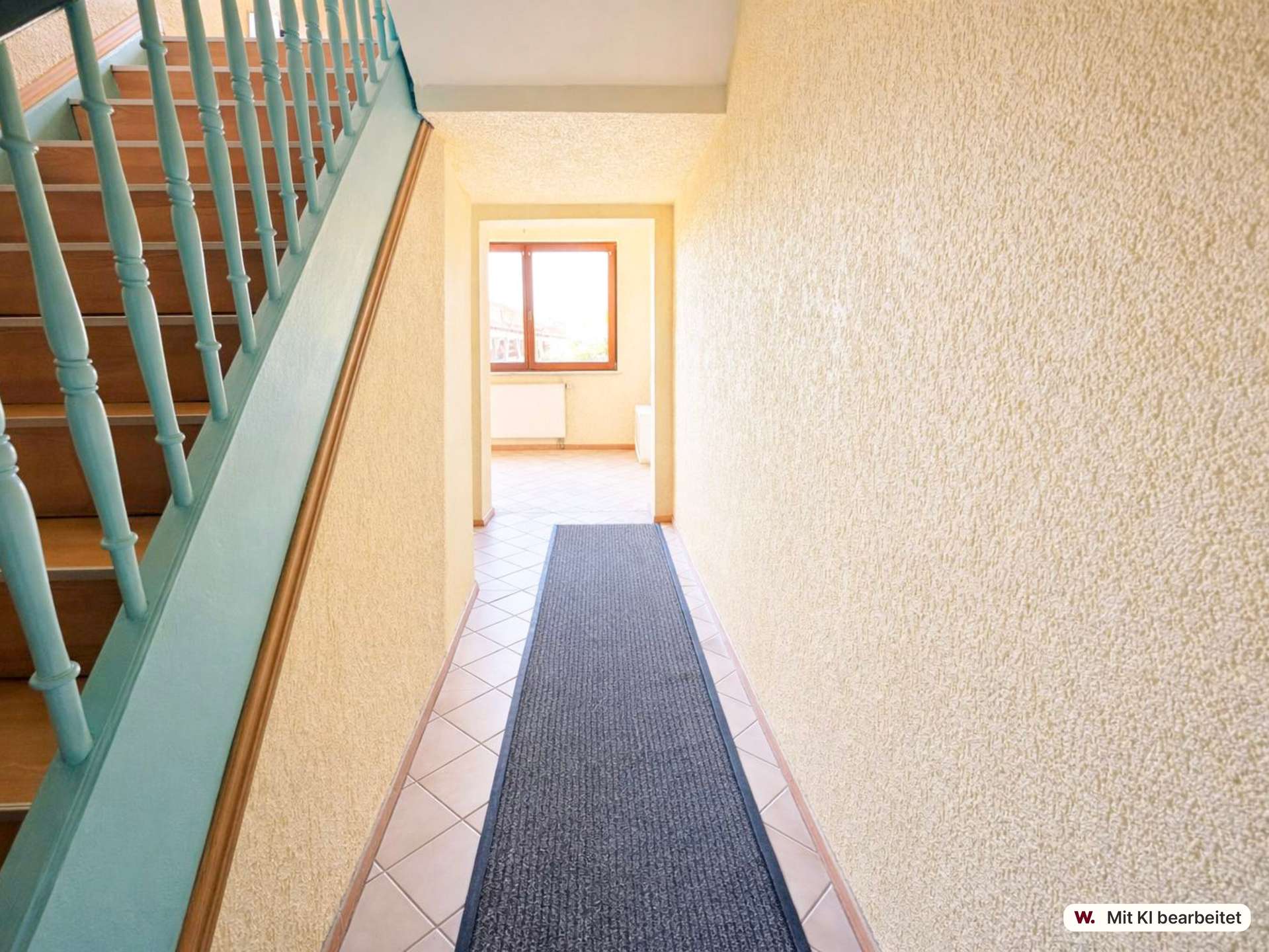 Indoor hallway and staircase with a turquoise railing on the left, beige textured walls, and a dark gray rug along tiled floor toward a sunlit doorway at the end.