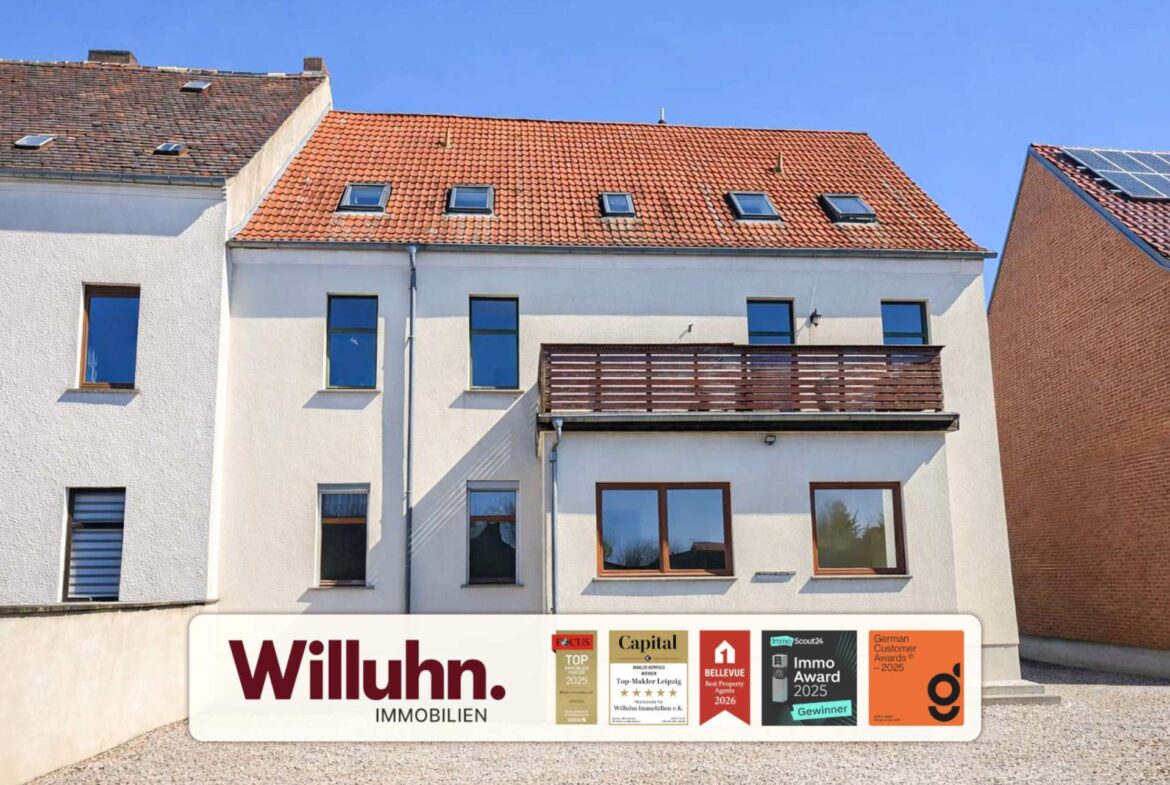 White apartment building with a red tile roof and skylights; a large Willuhn Immobilien banner with award logos is at the bottom.