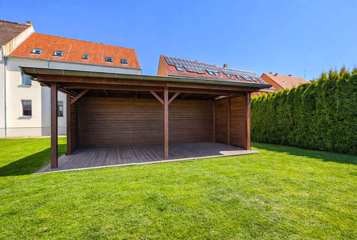 Wooden carport in a grassy backyard, with a house and red-tiled roof and solar panels on the neighboring roofs under a blue sky.