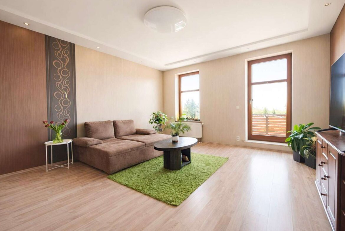 Living room with a brown sectional sofa, round coffee table, and a bright green rug near large windows and plants.