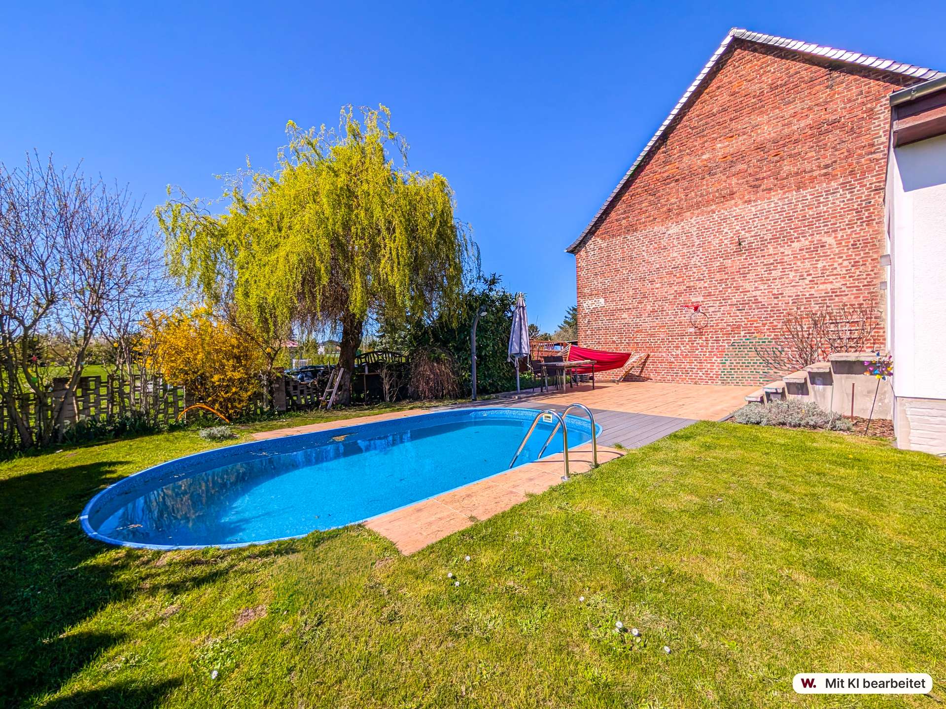 Backyard with a blue swimming pool, brick house wall, and patio furniture under a clear blue sky.