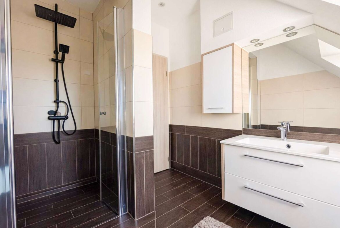 Modern bathroom with a glass shower enclosure, black wall-mounted shower system, white vanity, and beige-brown tiled walls and floor.