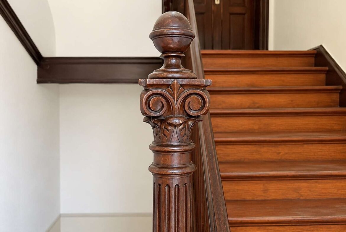 Ornate carved wooden stair newel post at the bottom of a polished hardwood staircase with a round finial.