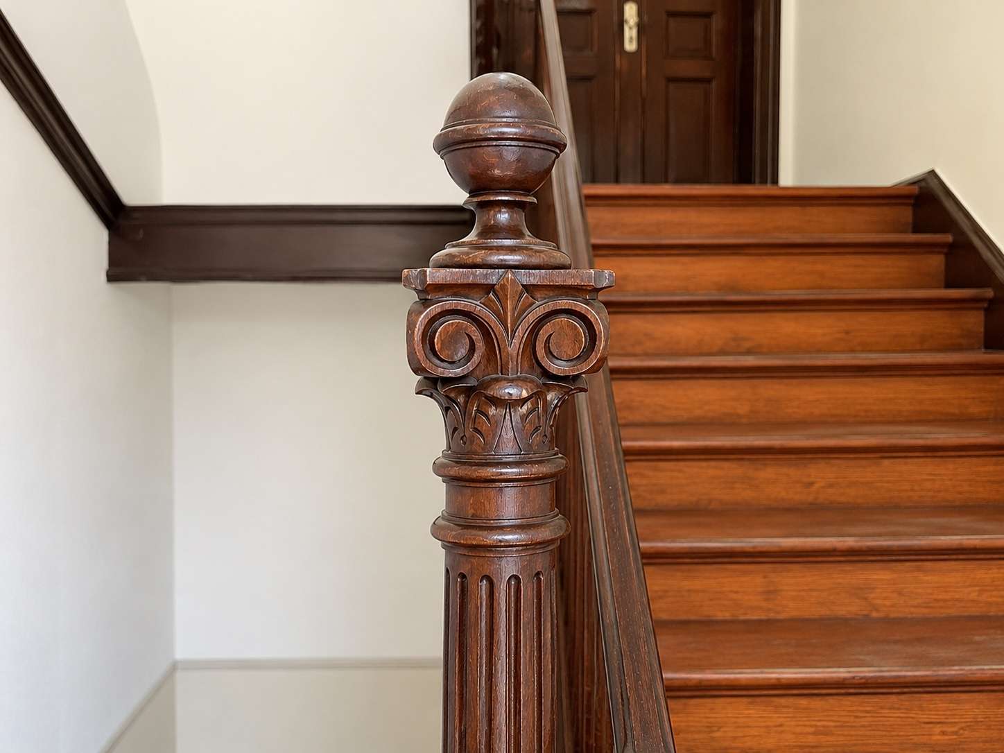 Ornate carved wooden stair newel post at the bottom of a polished hardwood staircase with a round finial.