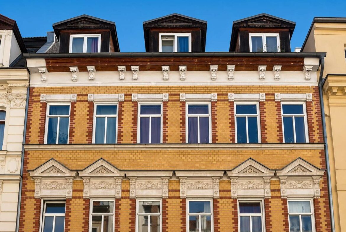 Brick building with ornate white decorative panels and tall windows under a clear blue sky; dormer roofs on top floor.