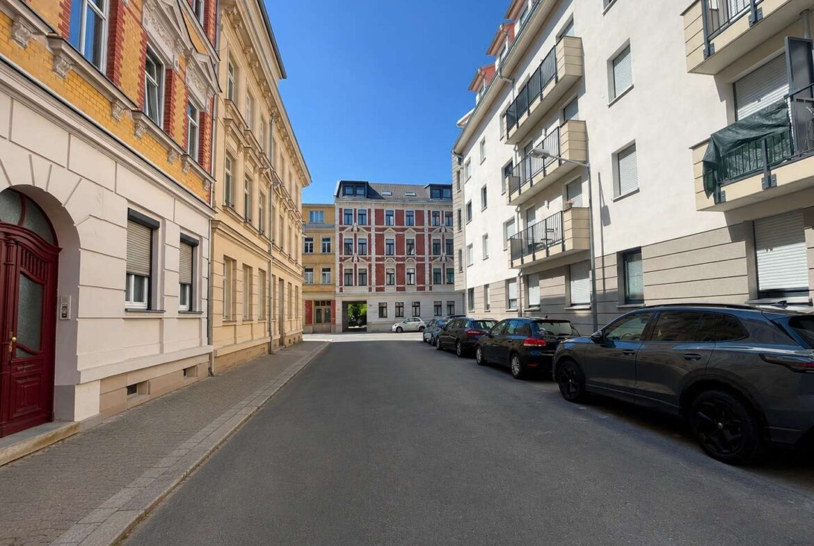 Residential street flanked by historic beige and red-brick buildings, parked cars along the curb, clear blue sky above.