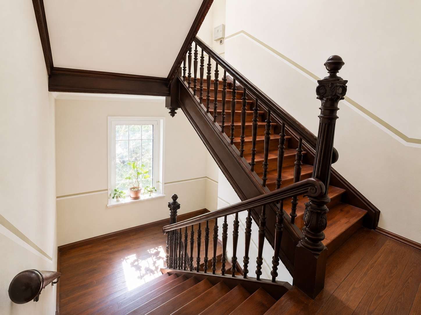 Formal dark-wood staircase with carved balusters and a large newel post, winding up between light walls in a bright entry hall with a window and potted plant on the sill.