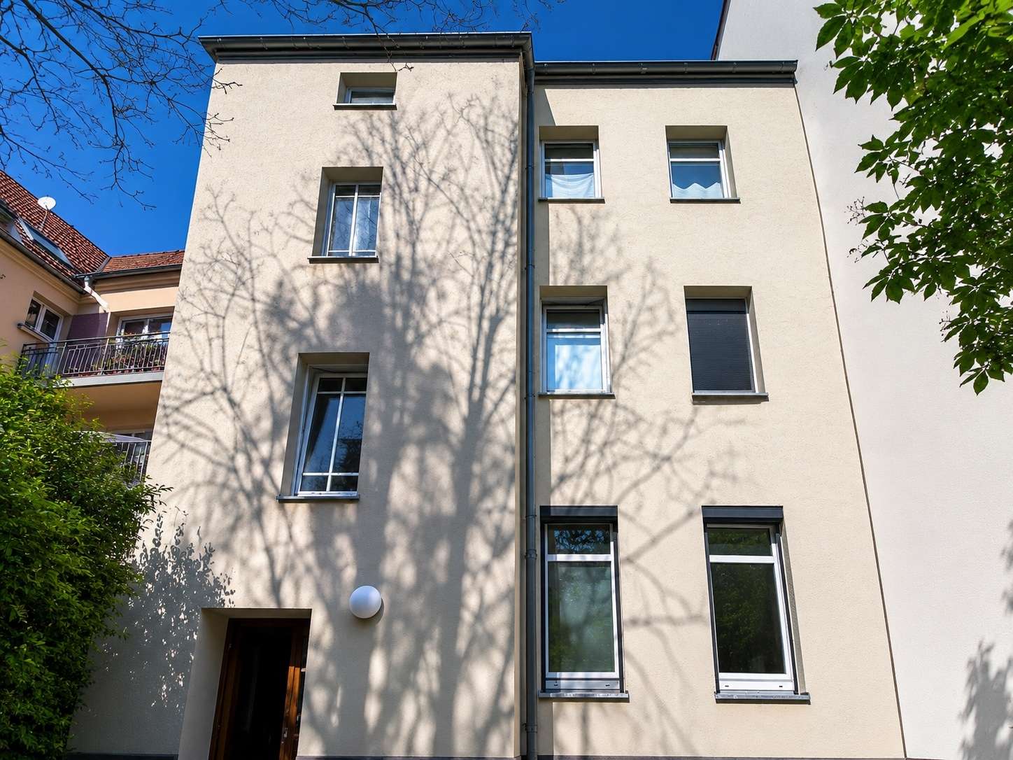 Facade of a beige, multi-story building with several rectangular windows and a dark downspout, under a bright blue sky; tree shadows cross the wall.