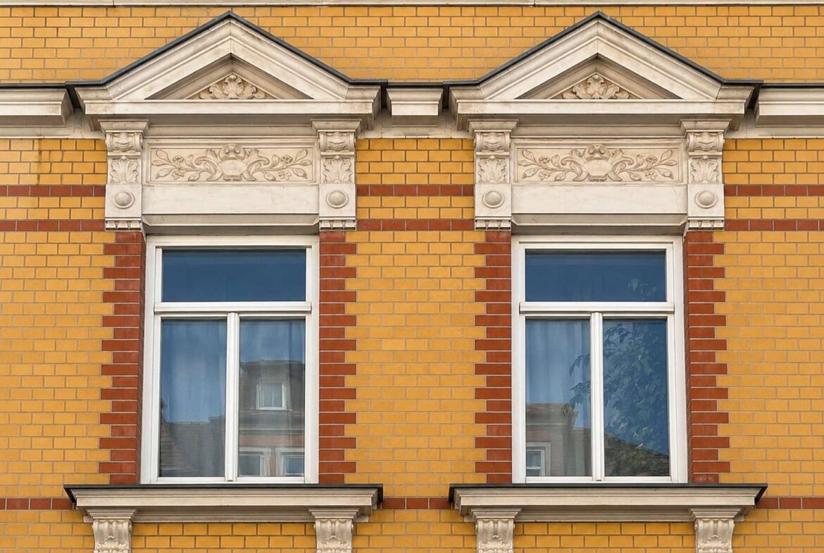 Two white framed sash windows set in a yellow brick facade with ornate stone pediments and decorative molding above each window