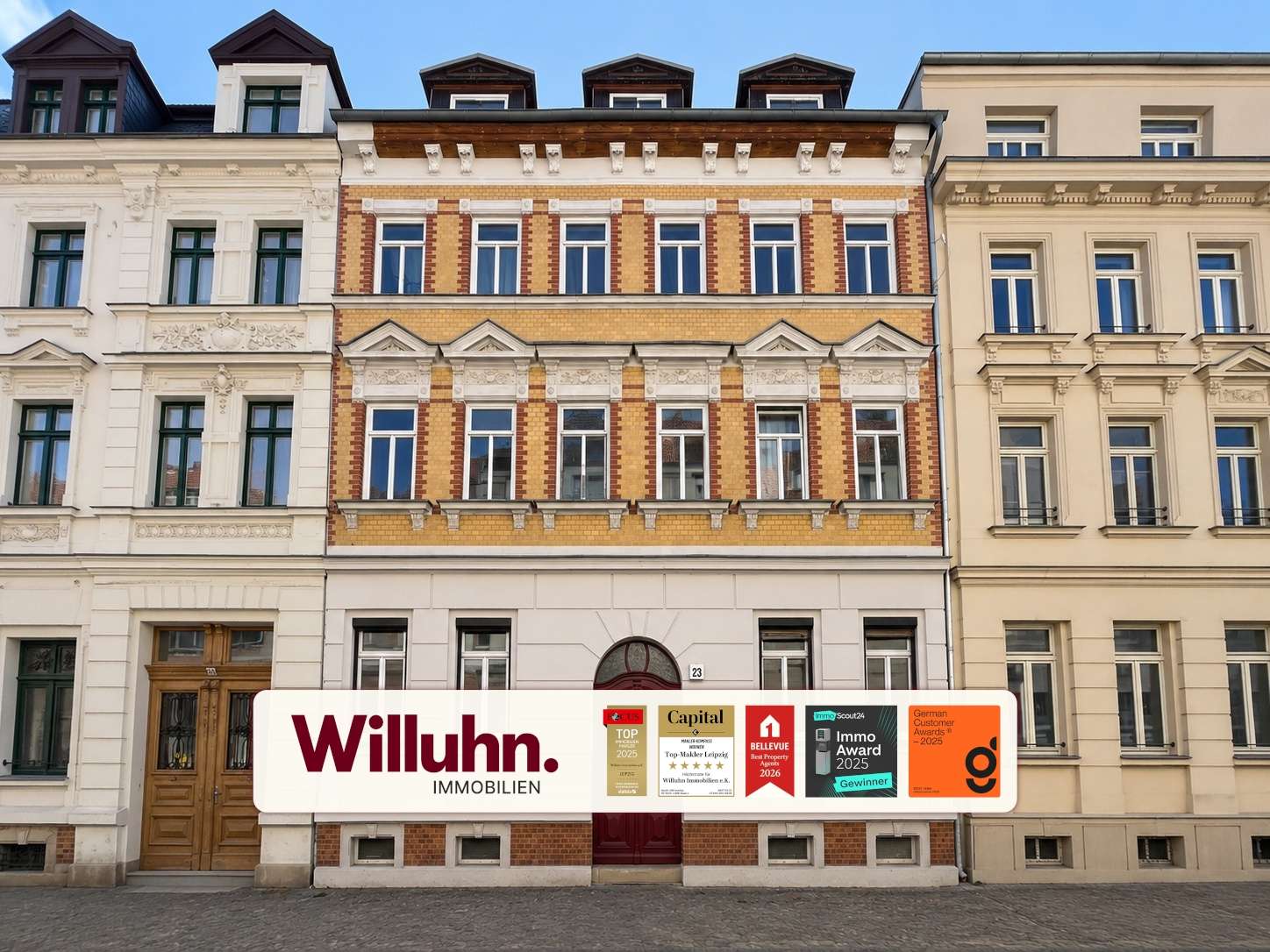 Facade of a row of historic buildings with a large 'Willuhn Immobilien' storefront sign and award plaques,