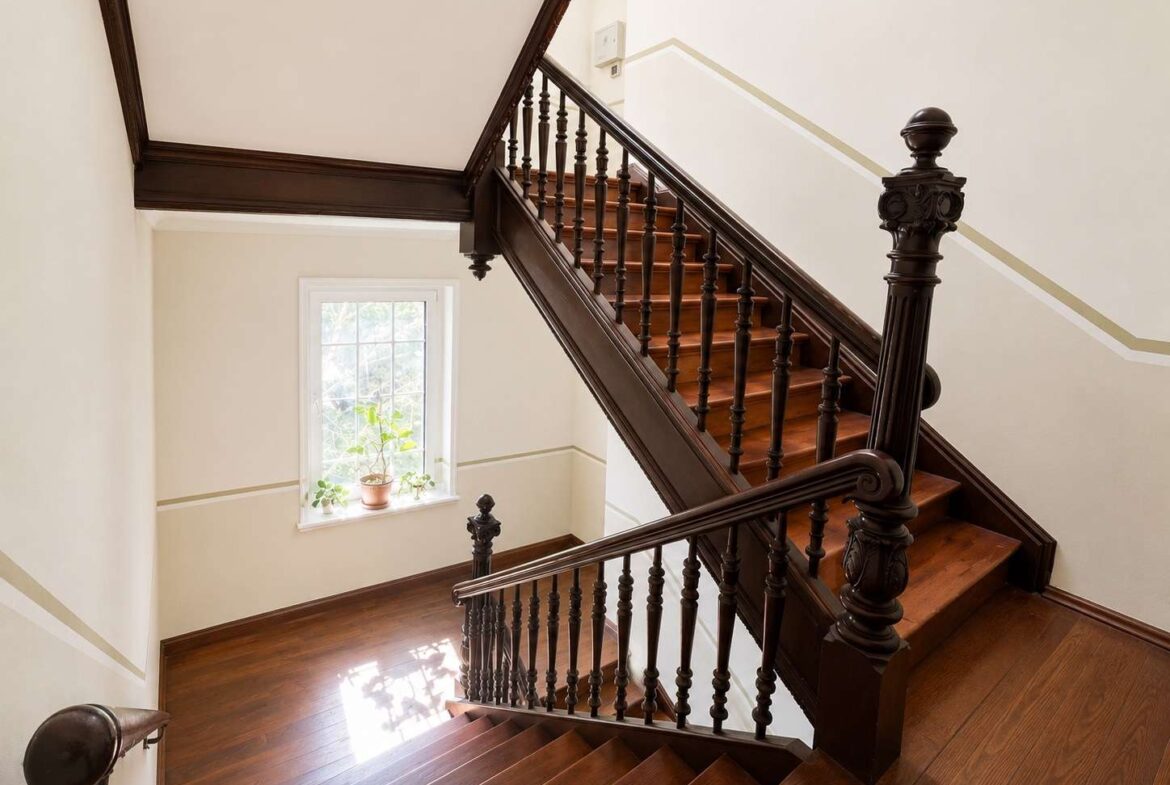 Indoor wooden staircase with dark carved banisters and polished steps, sunlight streaming in from a window with a potted plant on the sill.