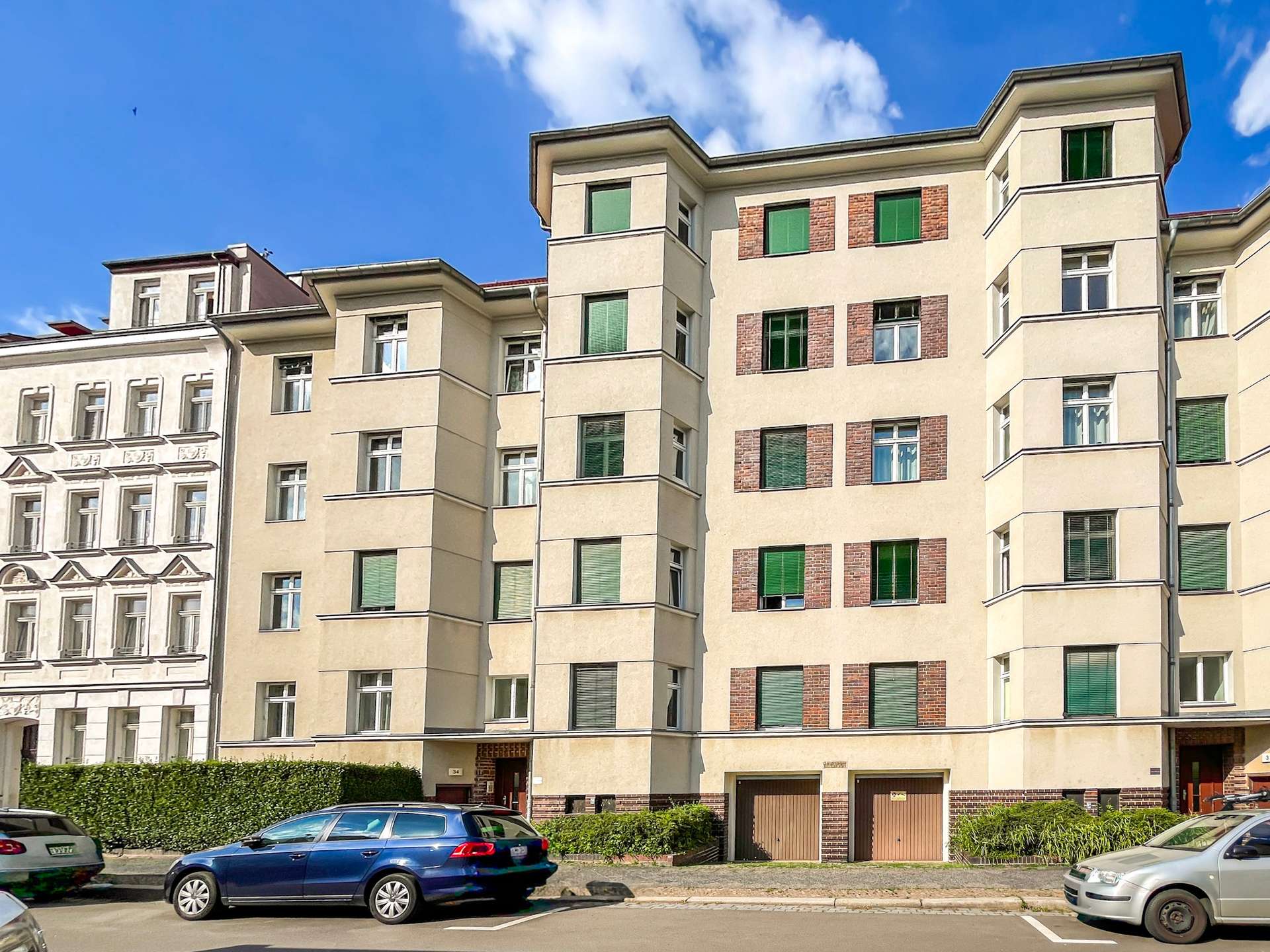Row of beige residential buildings with brick accents and green-framed windows, parked cars in front.