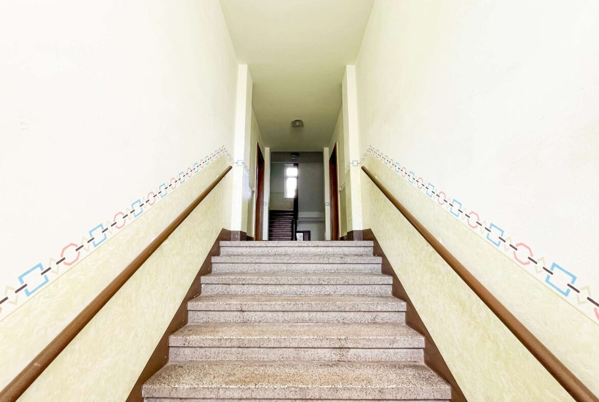 Interior stairwell with pale yellow walls, brown handrails, and a geometric tile border, viewed from the bottom looking up toward doors at the top.