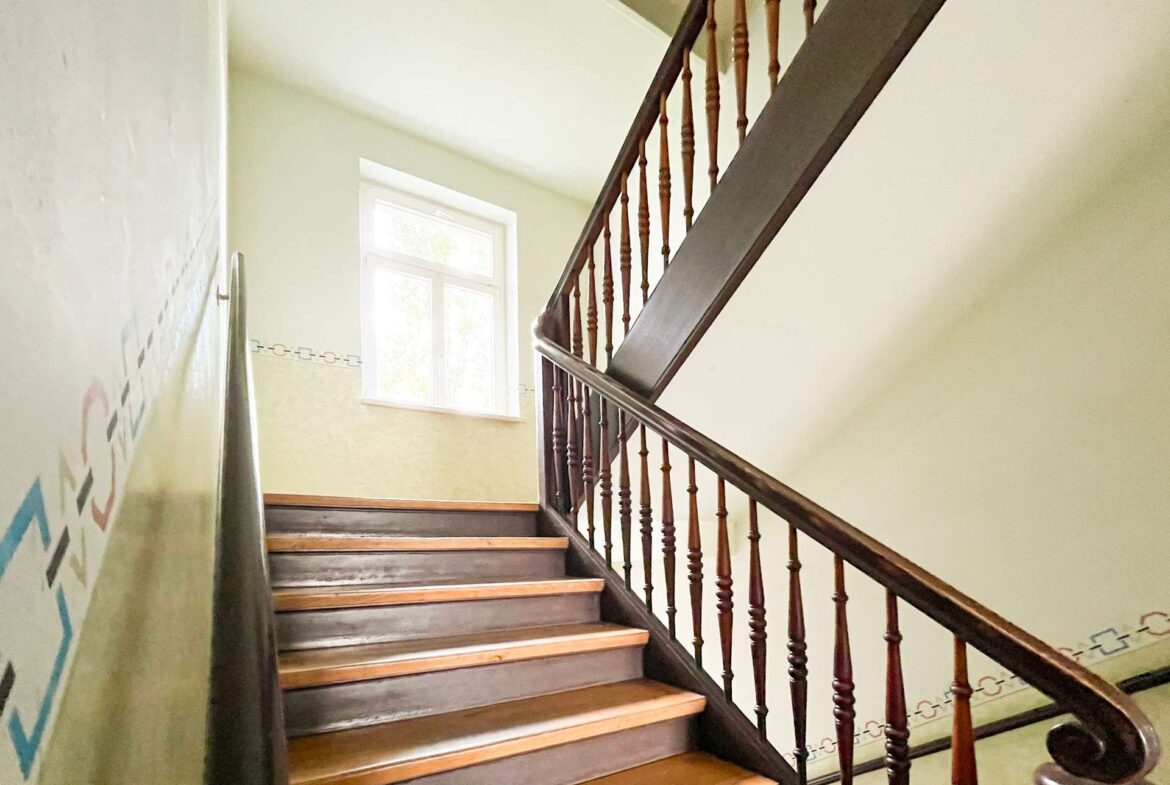 Interior stairwell with a dark wooden handrail and spindled banisters, illuminated by a window at the landing.