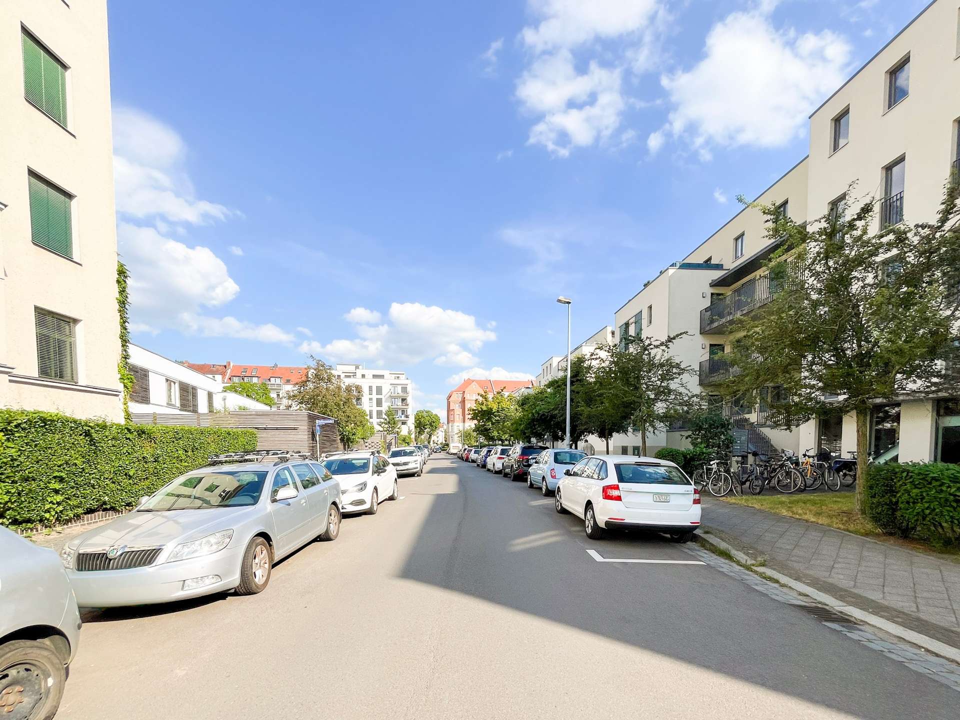 Residential street lined with parked cars on both sides and modern apartment buildings, bicycles on the right, blue sky above.