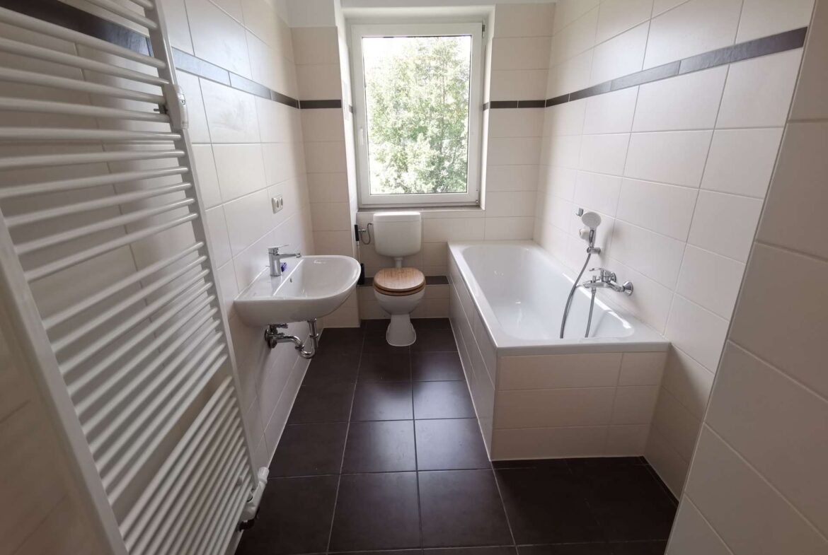 Bright white-tiled bathroom with a window above the toilet; sink to the left and a bathtub to the right, plus a wall-mounted white towel radiator on the far left.
