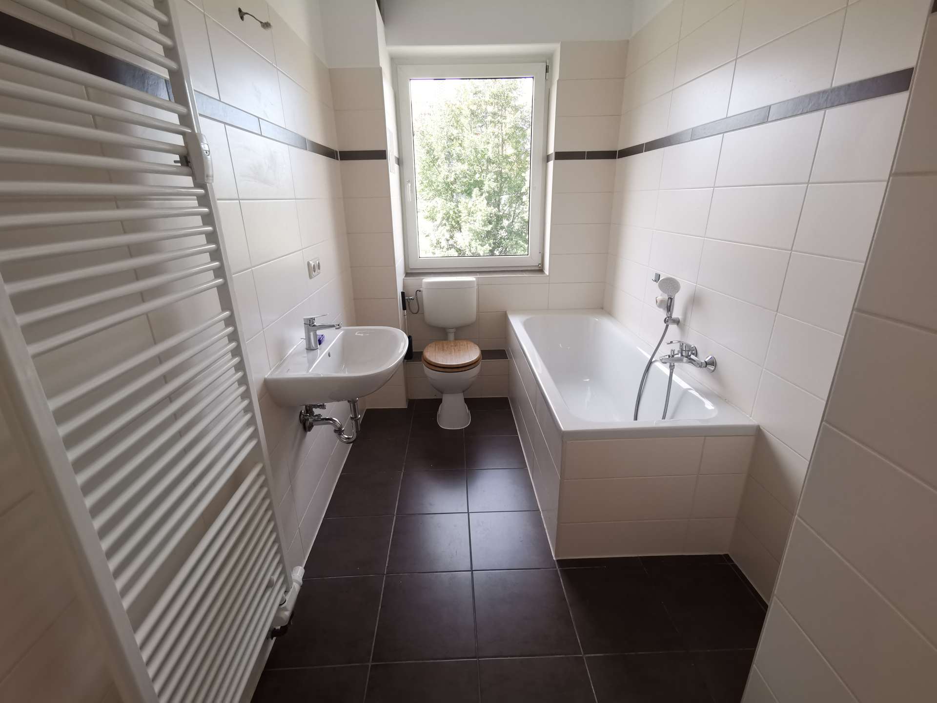 Bright white-tiled bathroom with a window above the toilet; sink to the left and a bathtub to the right, plus a wall-mounted white towel radiator on the far left.