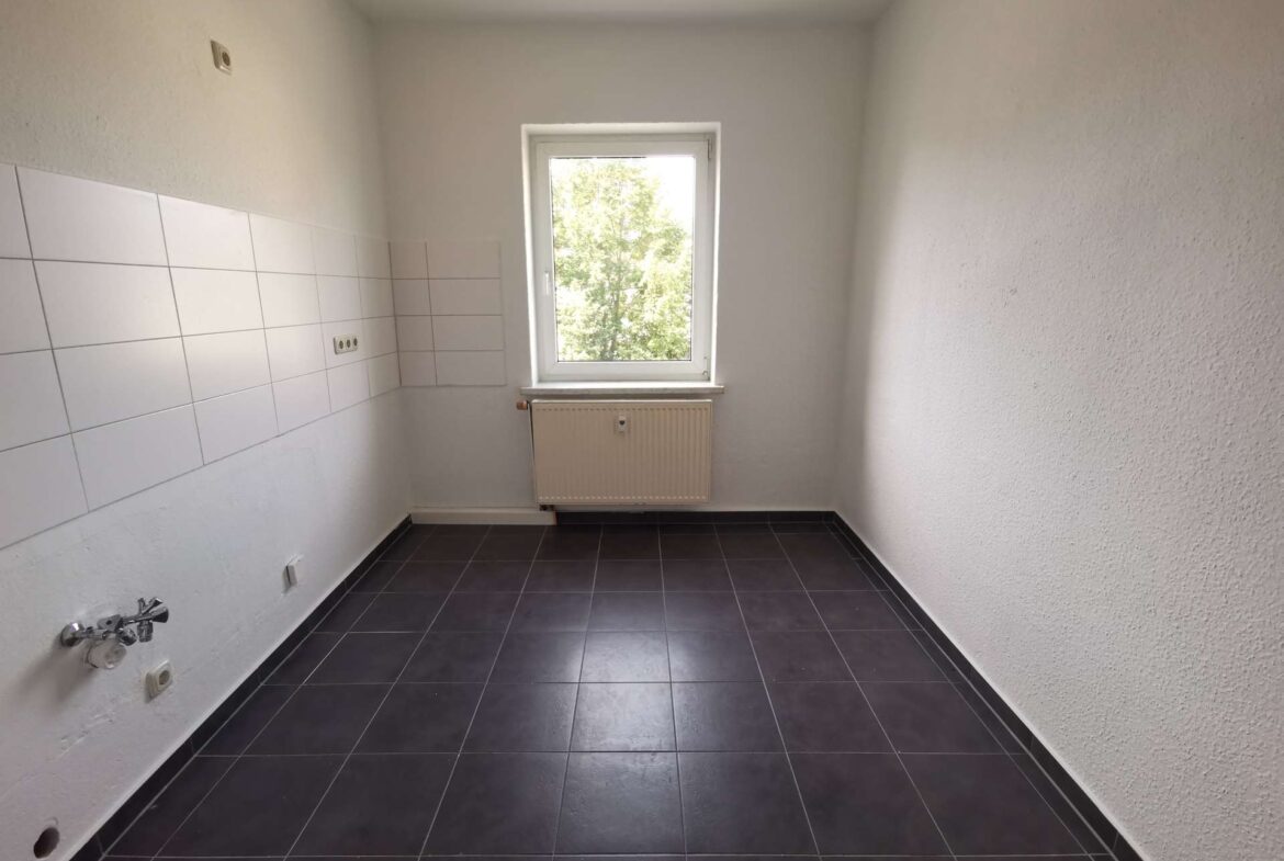 Empty kitchen with white tiles along the left wall, window above a radiator, and dark floor tiles.