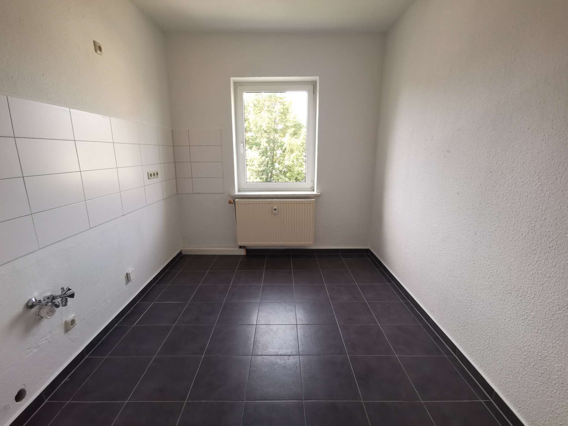 Empty kitchen with white tiles along the left wall, window above a radiator, and dark floor tiles.