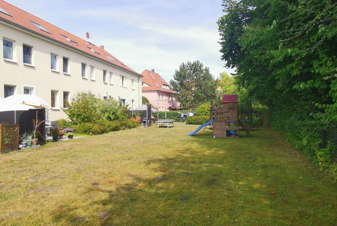 Apartment building with red-tiled roof along a sunny courtyard garden and a children's playset in the foreground under a blue sky.