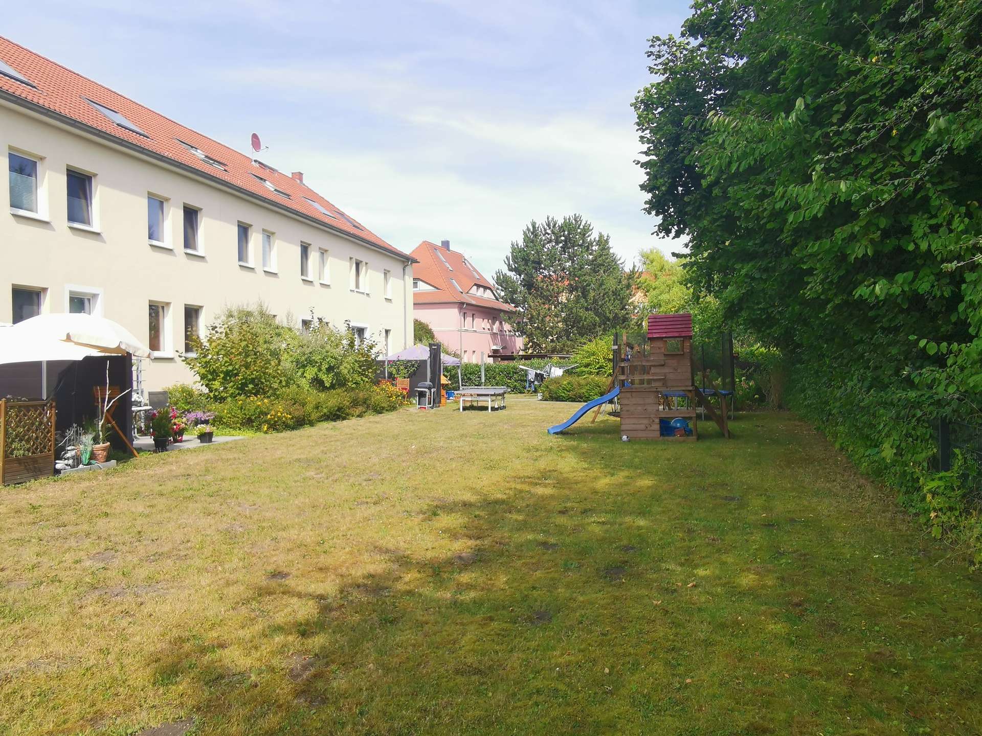 Apartment building with red-tiled roof along a sunny courtyard garden and a children's playset in the foreground under a blue sky.