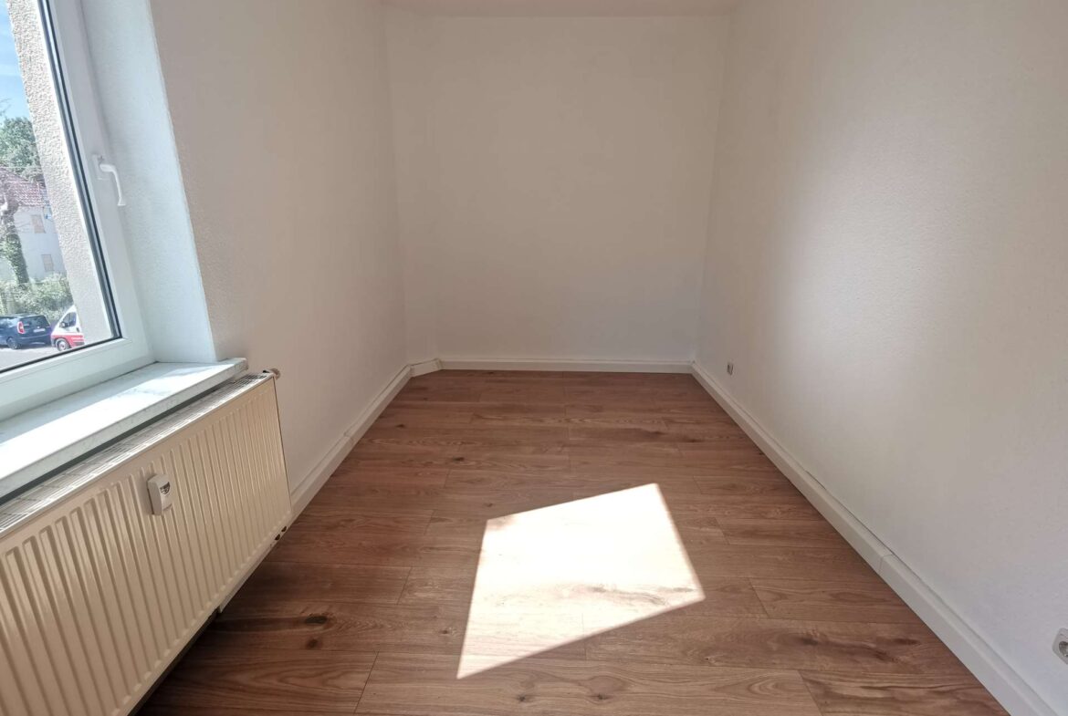 Empty, narrow room with white walls, a radiator under a large left-side window, and wooden floor bathed in sunlight.