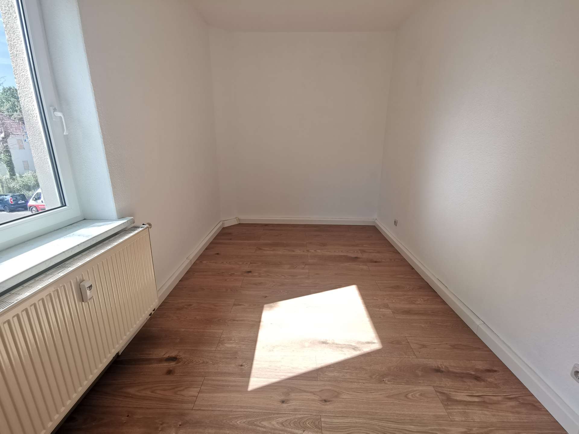 Empty, narrow room with white walls, a radiator under a large left-side window, and wooden floor bathed in sunlight.