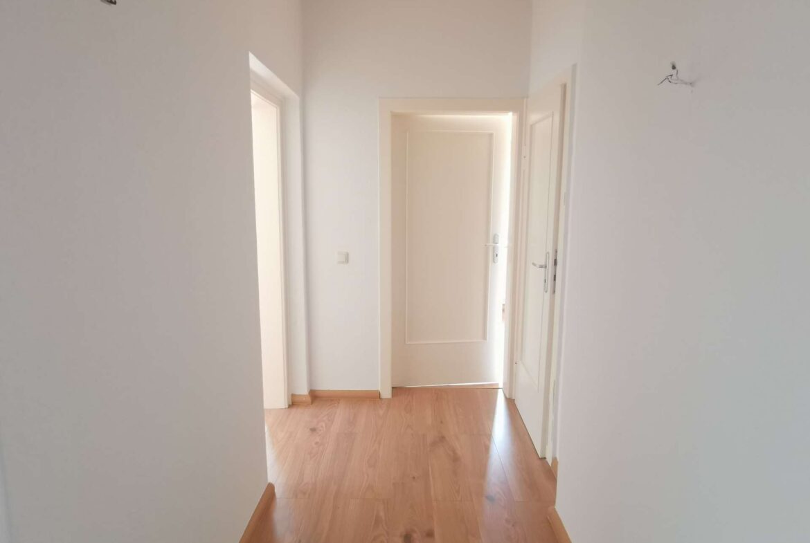 Bright white hallway with open doors on both sides and a wooden floor leading to a sunlit doorway at the end