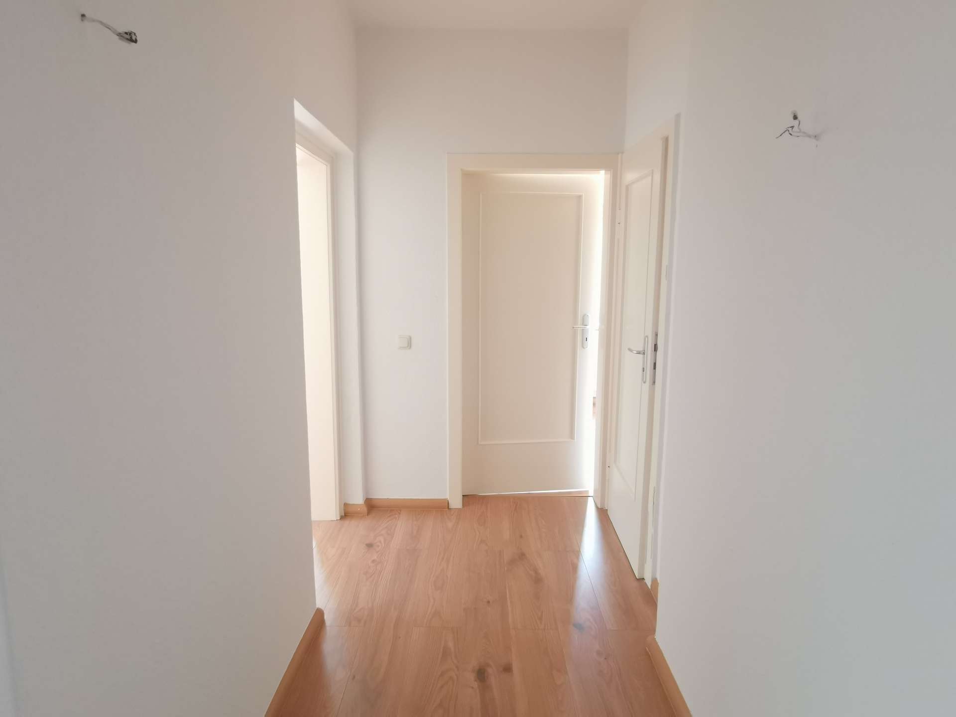 Bright white hallway with open doors on both sides and a wooden floor leading to a sunlit doorway at the end