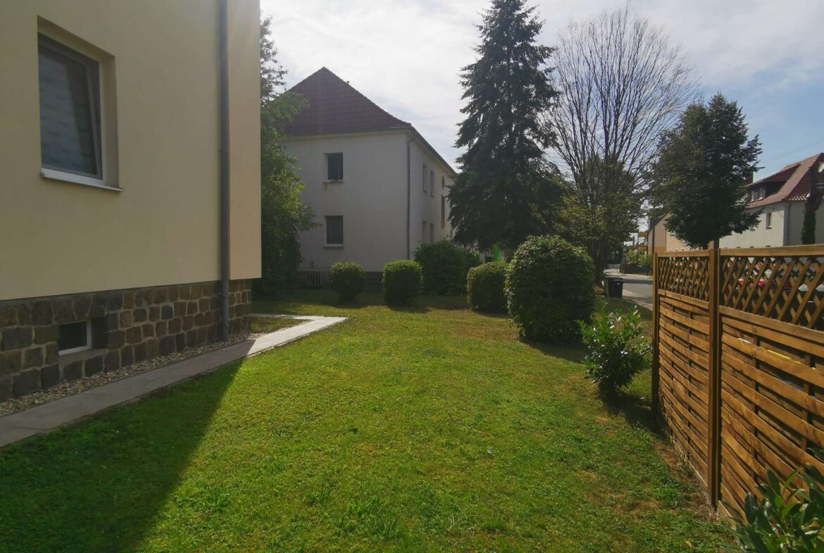 Suburban backyard with a beige building on the left, a green lawn, a paved path, and a wooden fence on the right.