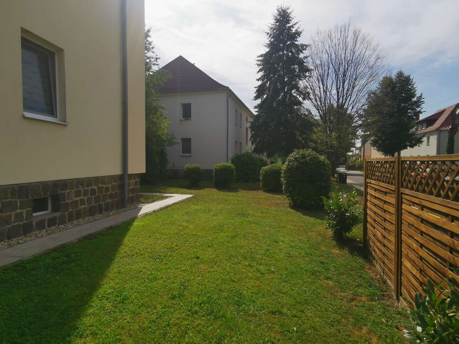 Suburban backyard with a beige building on the left, a green lawn, a paved path, and a wooden fence on the right.