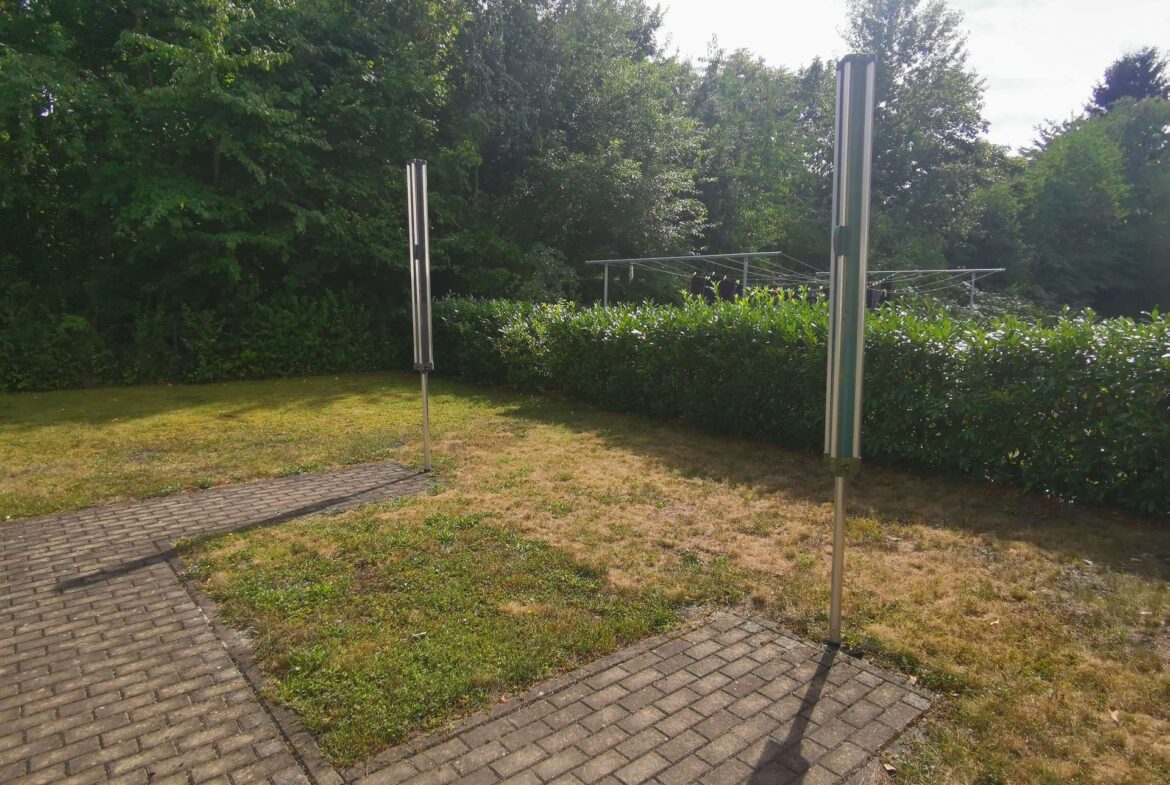 A suburban yard with a rotary clothesline behind a green hedge and two tall metal posts, under sunny sky.