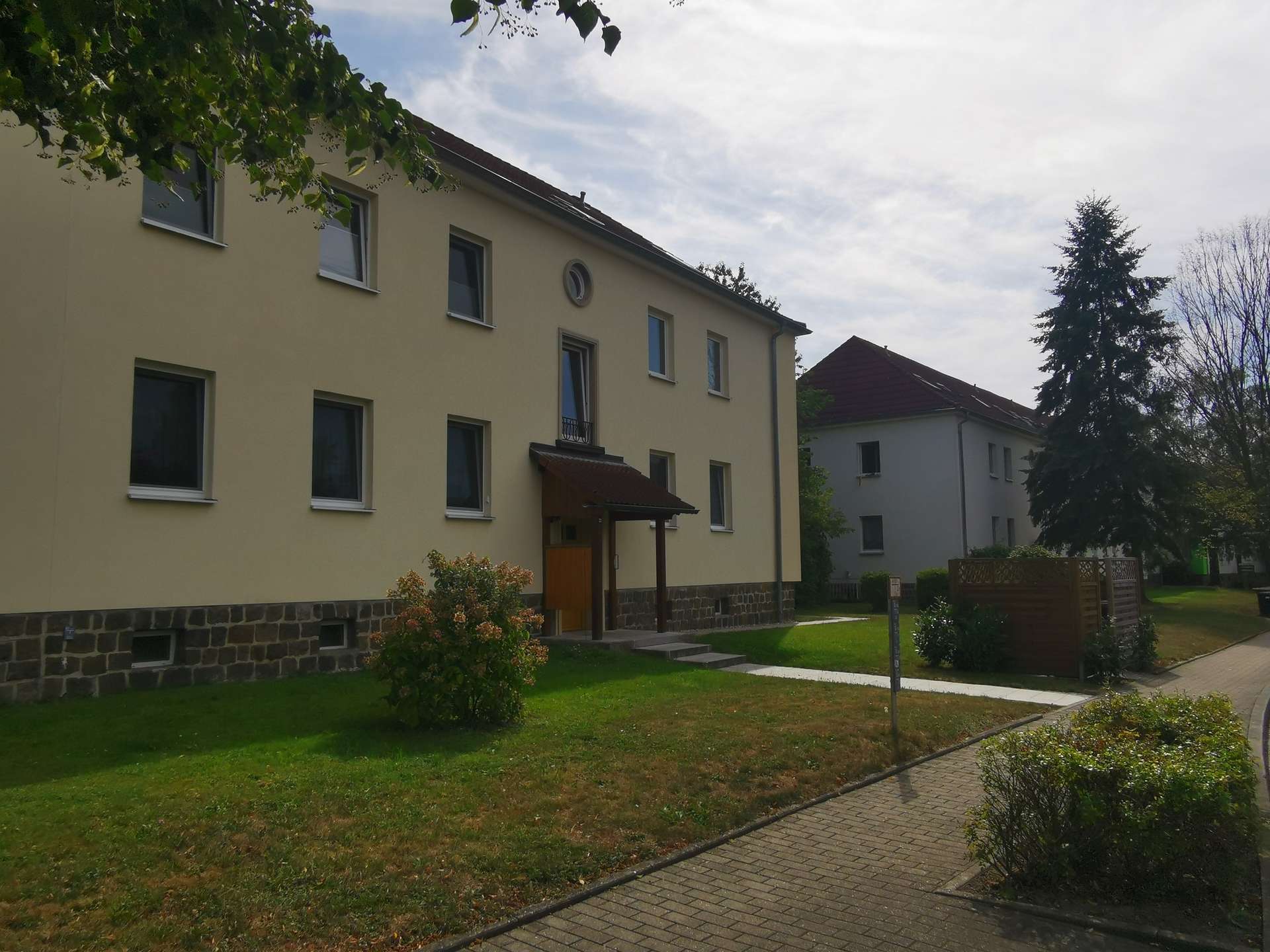 Front view of a beige, multi-story apartment building with numerous windows and a small entrance, set in a green lawn and trees.