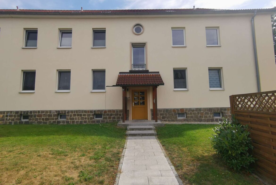 Beige apartment building with a central wooden door under a small red-tiled awning, a circular window above, and a paved path leading to the entrance.