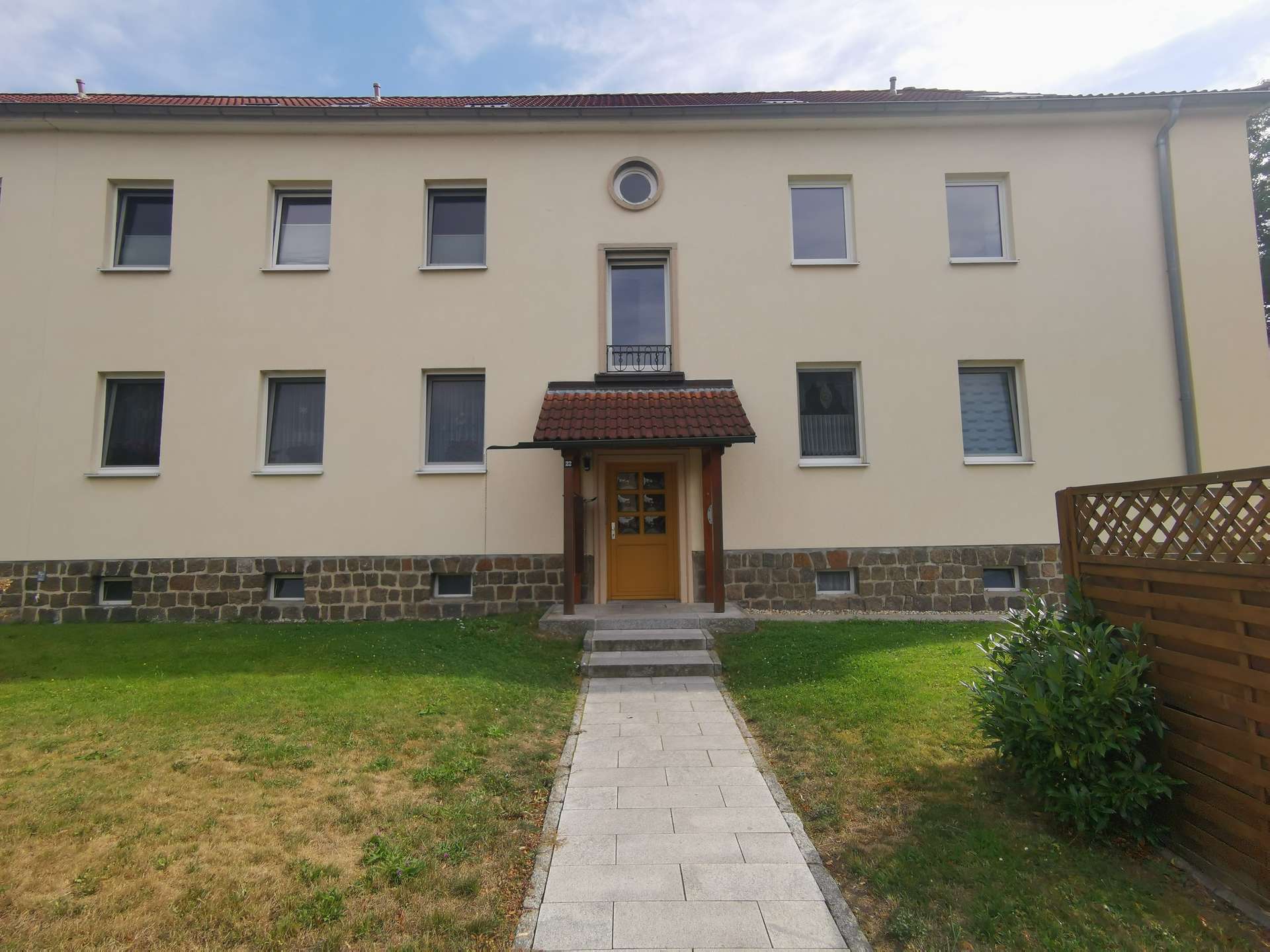 Beige apartment building with a central wooden door under a small red-tiled awning, a circular window above, and a paved path leading to the entrance.