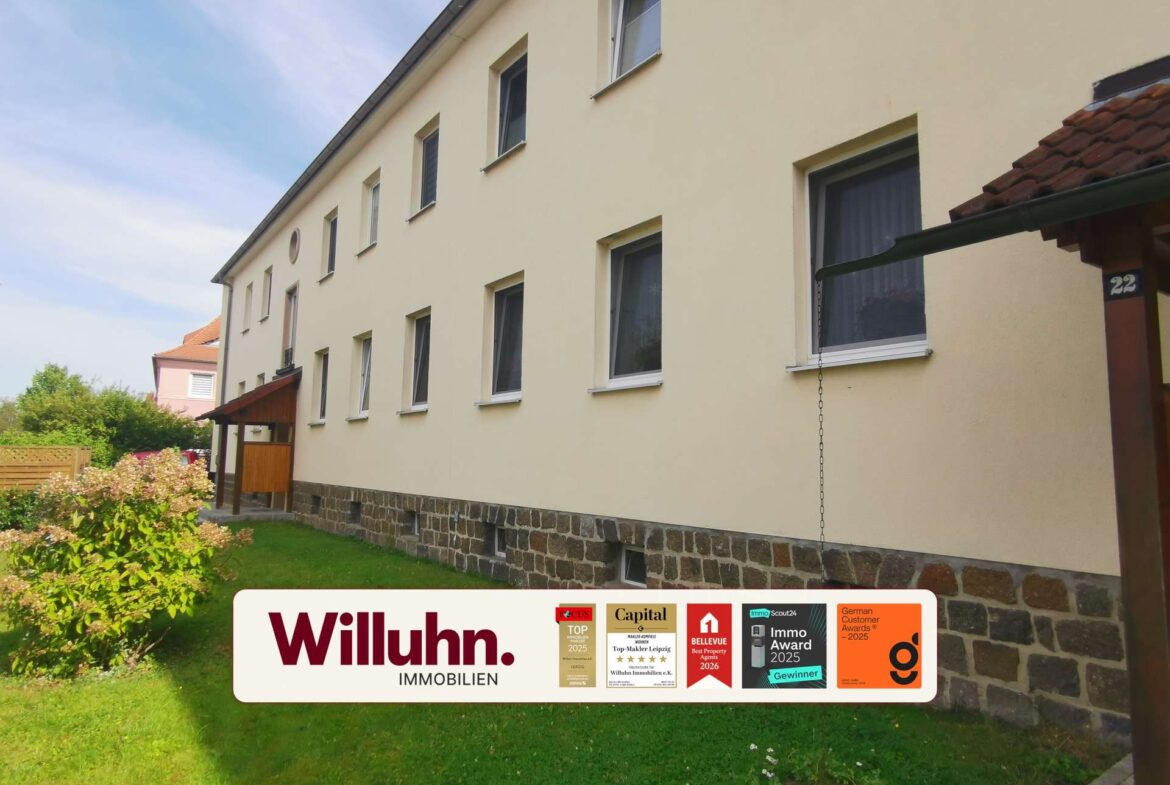 Exterior view of a light-yellow apartment building with multiple windows and a stone foundation, featuring a Willuhn Immobilien sign with awards.