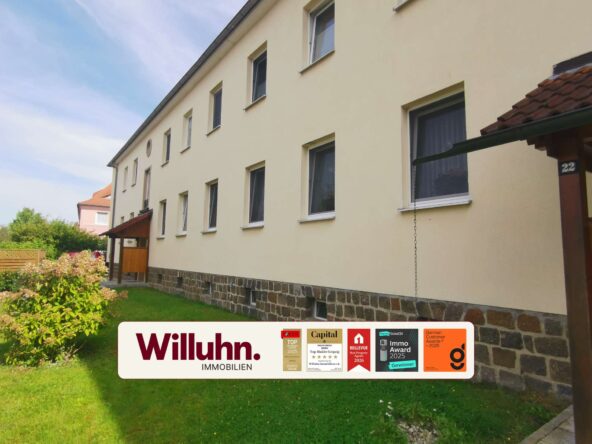 Exterior view of a light-yellow apartment building with multiple windows and a stone foundation, featuring a Willuhn Immobilien sign with awards.