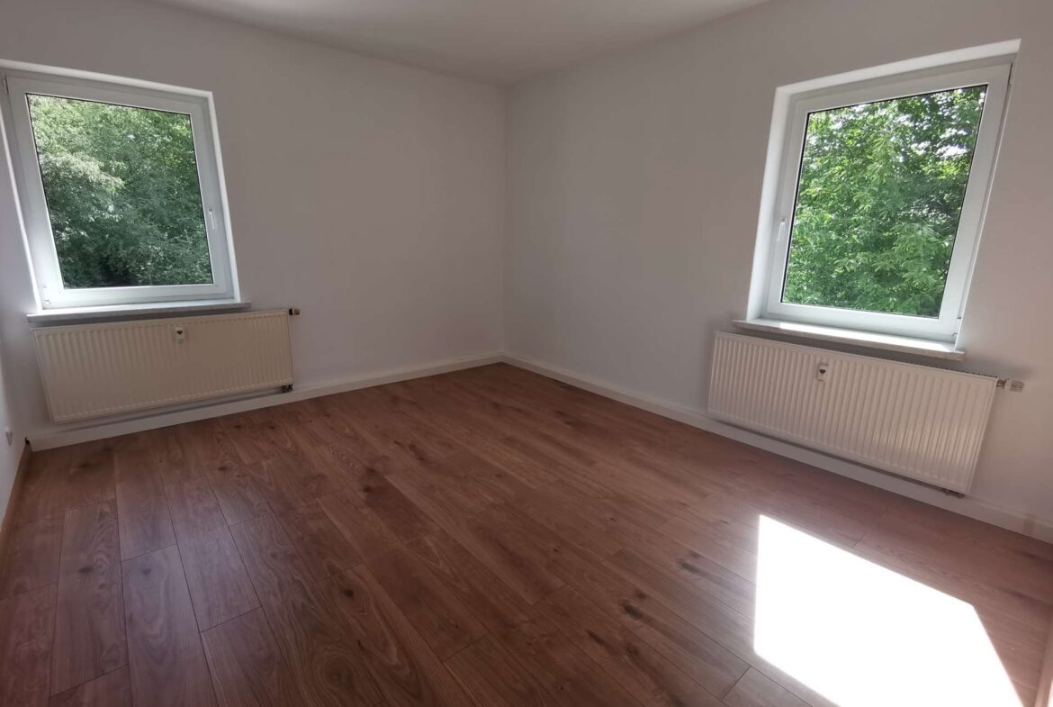 Empty room with two large windows, white walls, and wood-look flooring; radiators under each window and a view of green trees outside.