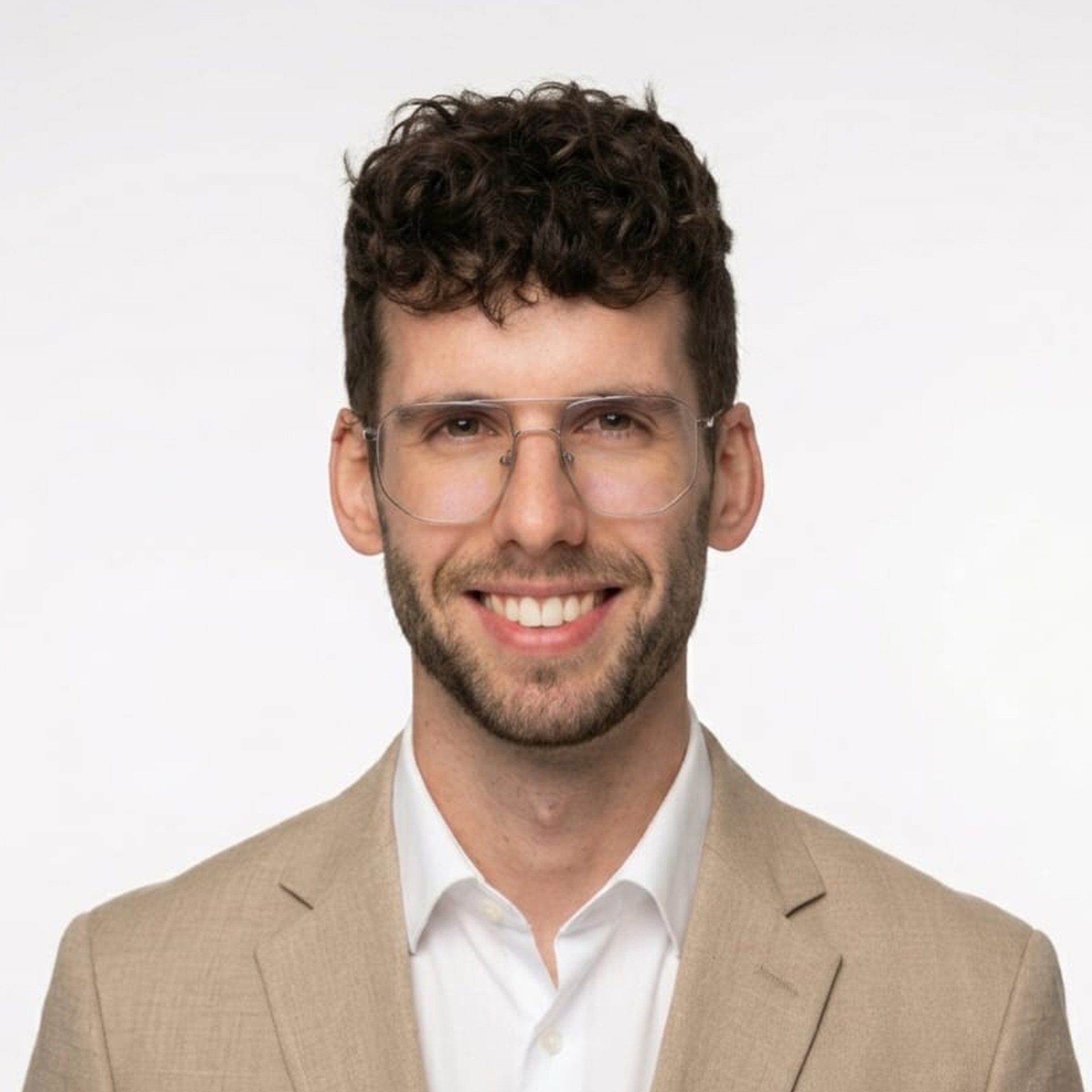 Young man in a beige blazer and white shirt wearing clear safety glasses, smiling at the camera against a light background.