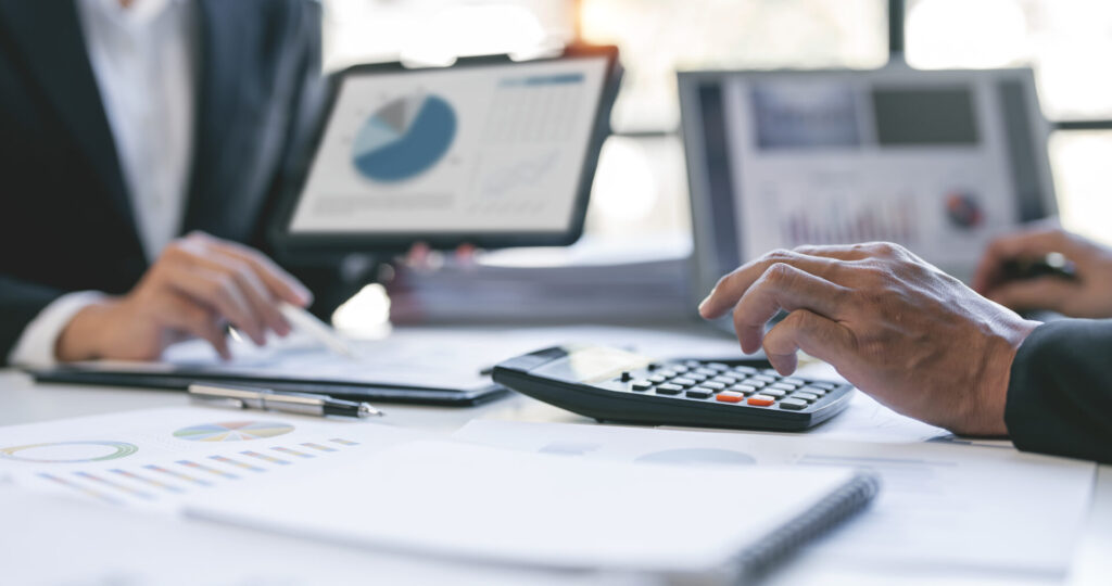 Two professionals analyze financial charts at a desk, using a calculator and documents on a blurred office table.